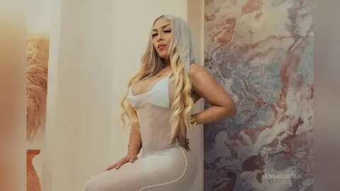 Glamorous blonde woman in a sheer white mesh dress posing against a marble wall in a bright, elegant setting.