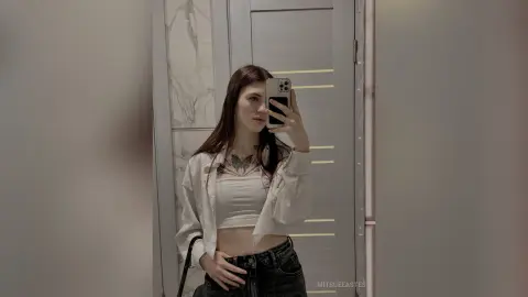 MitsueEastes poses for a mirror selfie wearing a white crop top and black jeans, showing off her long dark hair and chest tattoo.