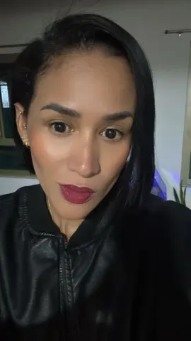 Smiling woman with dark hair wearing a black leather jacket in a dimly lit indoor setting.