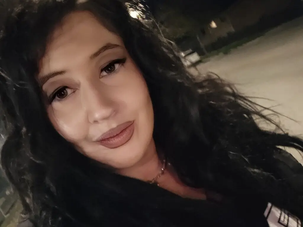 Smiling woman with long wavy dark hair and glamorous makeup posing outdoors at night.