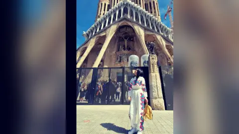 Stylish woman in white jumpsuit standing before the Sagrada Familia cathedral in sunny Barcelona.