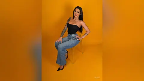 Smiling woman in a black tube top and blue jeans sitting on a chair against a vibrant yellow background.