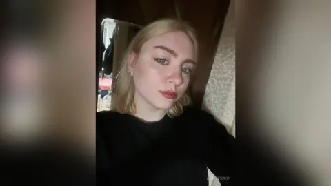 AnnyFraid looks into the camera with blonde hair and red lipstick, wearing a black shirt in a dimly lit room.