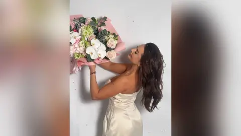 Woman in a white satin dress holding a large bouquet of pink and white flowers against a white background.