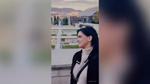 Smiling young woman in a white turtleneck and black jacket standing by a city fountain at sunset.
