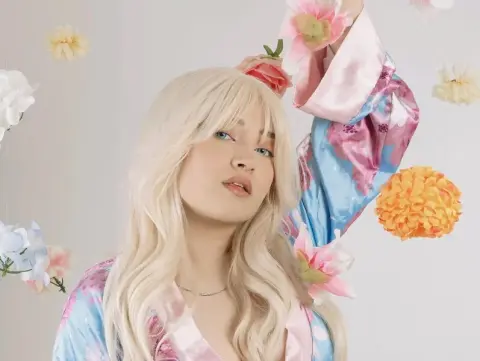 Blonde woman in a blue floral satin robe posing amidst colorful paper flowers in a bright, dreamy setting.