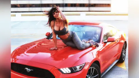 DrellaWolf — Athletic woman in grey gym wear posing confidently on a red Ford Mustang in a sunny parking lot.