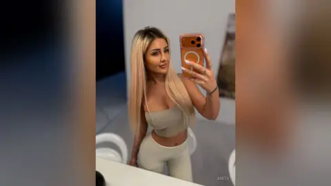 Anita poses for a mirror selfie wearing a light tan sports bra and matching leggings, letting her long blonde hair hang down her shoulders.