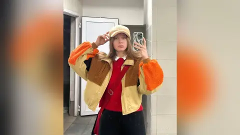 Young woman in a trendy beige and orange fleece jacket and red sweater posing in a mirror for a casual selfie.