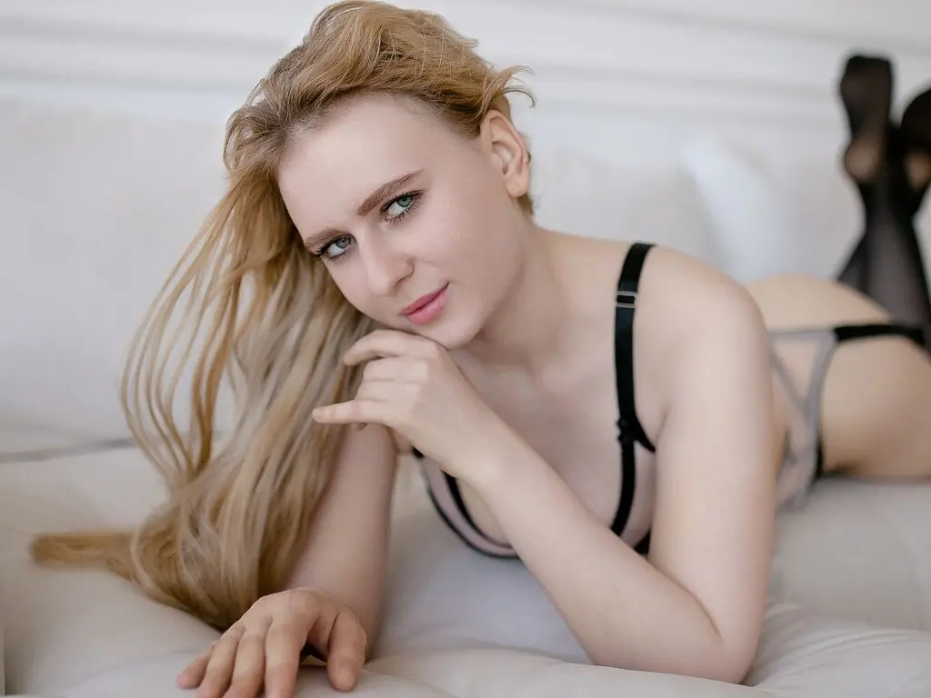ShinyLissa lies on a white bed wearing a black and grey lace bra and black stockings, looking at the camera with her blonde hair falling over her shoulder.