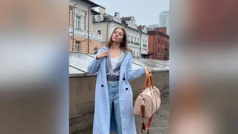 Young woman in a long light blue coat and jeans holding a designer handbag on a city street.