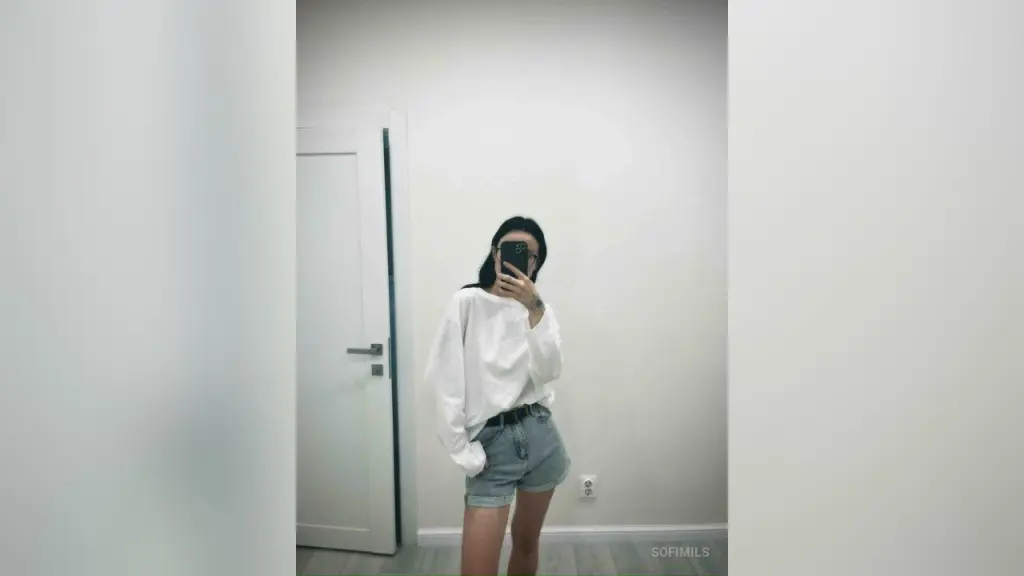 SofiMils β SofiMils stands against a white wall wearing a white long-sleeve shirt and denim shorts while taking a mirror selfie.