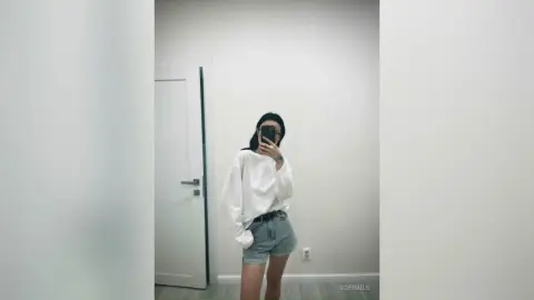 Young woman in a white oversized shirt and denim shorts taking a mirror selfie in a minimalist room.