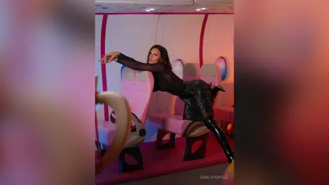 Stylish woman in a black outfit leaning back in a pink airplane cabin with a moody, cinematic vibe.