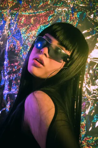 Edgy woman in black outfit and futuristic sunglasses posing against a colorful holographic glitter background.