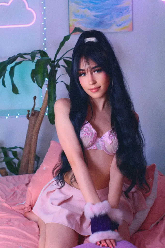 ThaliaZafirr — Young woman in pink lingerie and fuzzy gloves sitting on a bed in a dreamy, neon-lit bedroom setting.