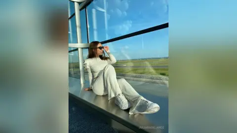 Stylish woman in white loungewear sitting by a large window overlooking a sunny landscape.