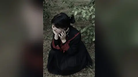 Melancholic woman in a vintage black and red dress sitting in a moody, overgrown outdoor garden setting.