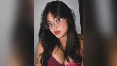 MadisonCarie wearing clear glasses and a maroon top, posing with long dark hair against a plain wall.