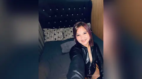 Smiling woman in a black leather jacket sitting on a bed with a dark tufted headboard in a cozy bedroom.