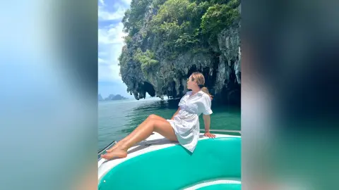 VictoriaWynn sits on the edge of a white boat, wearing a short white shirt, overlooking a tropical ocean and rocky cliffs.