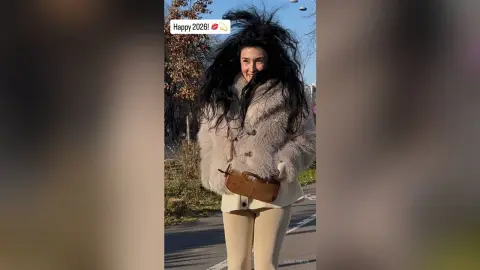 Smiling woman in a cozy beige faux fur coat and cream trousers standing outdoors on a sunny day.