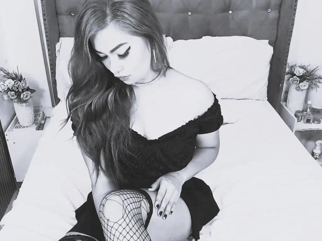 Moody black and white portrait of a woman in a black dress and fishnet tights sitting on a white bed.