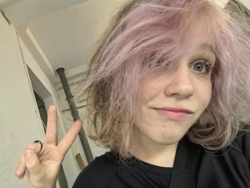 Young woman with messy pink hair making a peace sign in a casual indoor setting.