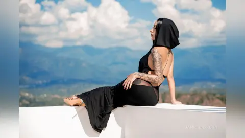 Tattooed woman in a black backless gown sitting on a white ledge against a scenic mountain landscape.