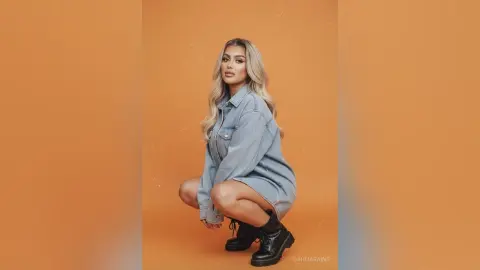 Fashionable woman in a denim shirt and black boots posing against a warm orange studio background.