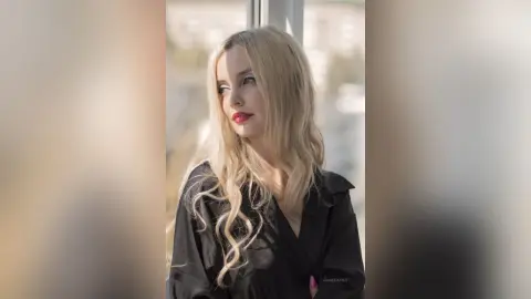 Blonde woman in a black blouse looking thoughtfully out a window in soft, natural sunlight.