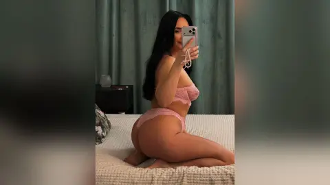Woman in pink lace lingerie taking a mirror selfie on a white bed in a cozy bedroom setting.