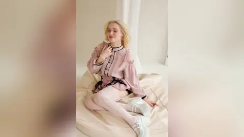 LynGoodhart sits on a white cushion wearing a pink ruffled blouse and white lace thigh-high stockings.