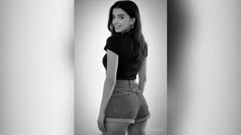 Young woman in a black t-shirt and denim shorts posing in a minimalist black and white studio setting.
