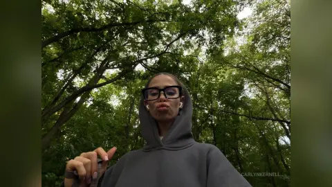 CarlynKernell wearing a grey hoodie and black glasses, posing outdoors under a canopy of green trees.