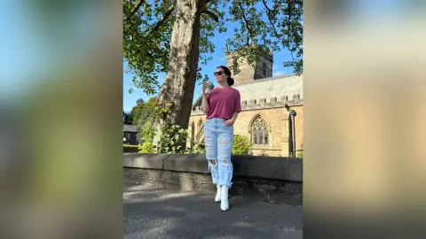 Woman in maroon t-shirt and ripped jeans holding a drink by a stone church under a sunny sky.
