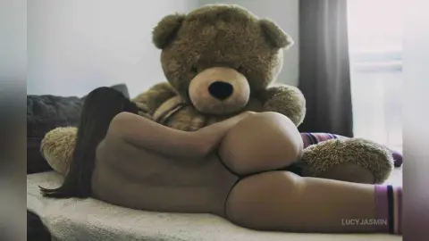 LucyJasmin — Nude woman in black lingerie lying on a bed next to a large brown teddy bear in a cozy bedroom setting.