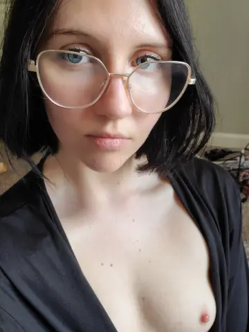 Close-up portrait of a young woman with short black hair and glasses wearing a black top in a soft indoor setting.