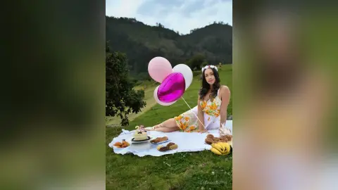 Young woman in a floral sundress enjoying a romantic picnic with pink balloons in a lush green meadow.