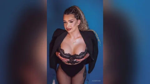 AvaBronX — AvaBronX poses in black lace lingerie and a black blazer against a blue background.