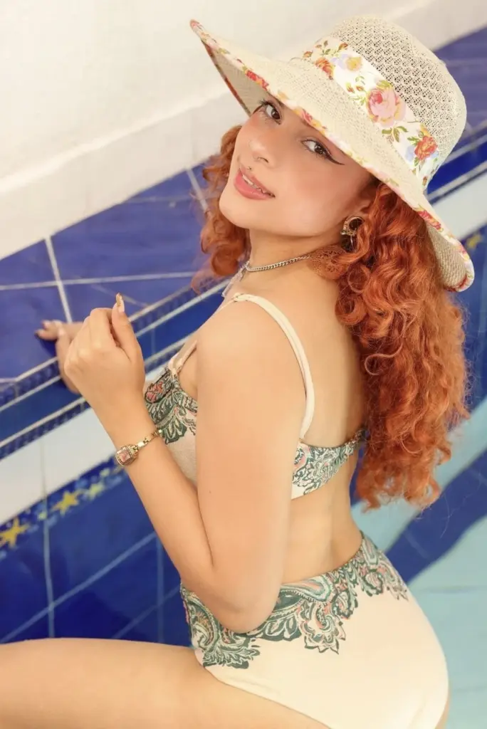 Historia poses in a white cut-out bodysuit and white hat, showing her red hair and curves while sitting by a pool or hot tub.
