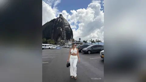 Woman in white crop top and trousers standing in a parking lot near a massive rock mountain under a cloudy sky.