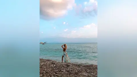 Blonde woman in a white floral maxi dress standing on a rocky beach under a dreamy sunset sky.