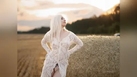Blonde woman in a white lace dress standing in a golden wheat field during a dreamy sunset.