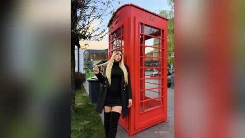 Blonde woman in a black dress and over-the-knee boots posing by a classic red British telephone booth.
