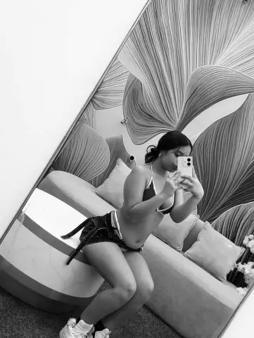 Young woman in a bikini and denim shorts taking a selfie in front of a floral patterned wall.