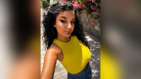 Smiling young woman with wavy dark hair wearing a bright yellow sleeveless top in a sunny outdoor garden setting.