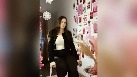 IngerMarzella poses in a black jacket and white top, sitting against a wall decorated with pink heart-themed photos and dried pampas grass.