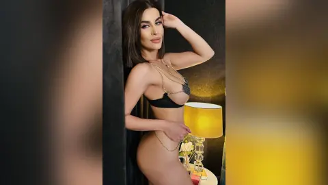 Glamorous woman in black bikini and gold chain jewelry posing in a moody, dimly lit indoor setting.