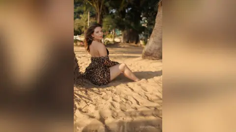 Young woman in a leopard print dress sitting on a sunny tropical beach near palm trees.